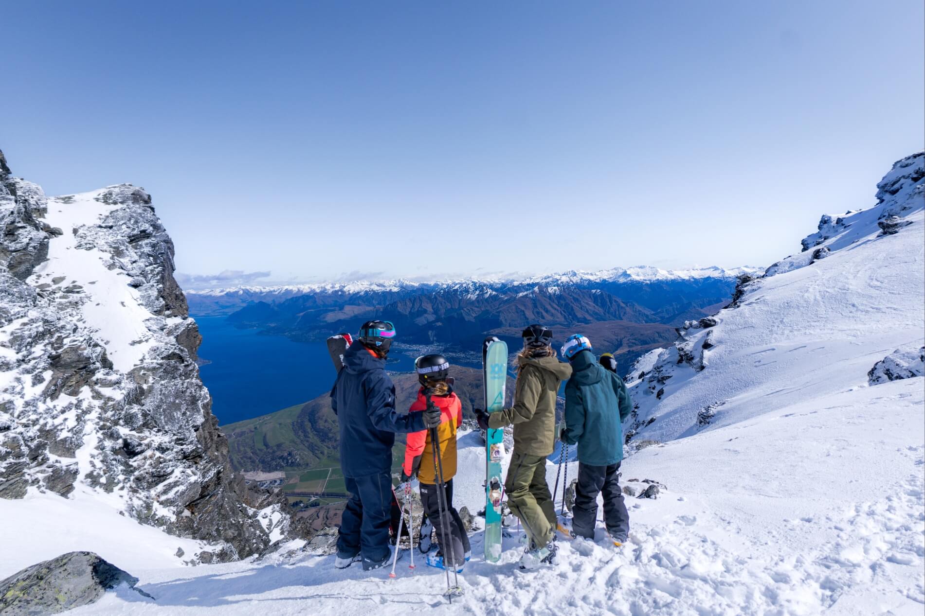 Four skiers standing on a snowy mountain peak overlooking a lake and distant mountains under a clear sky.