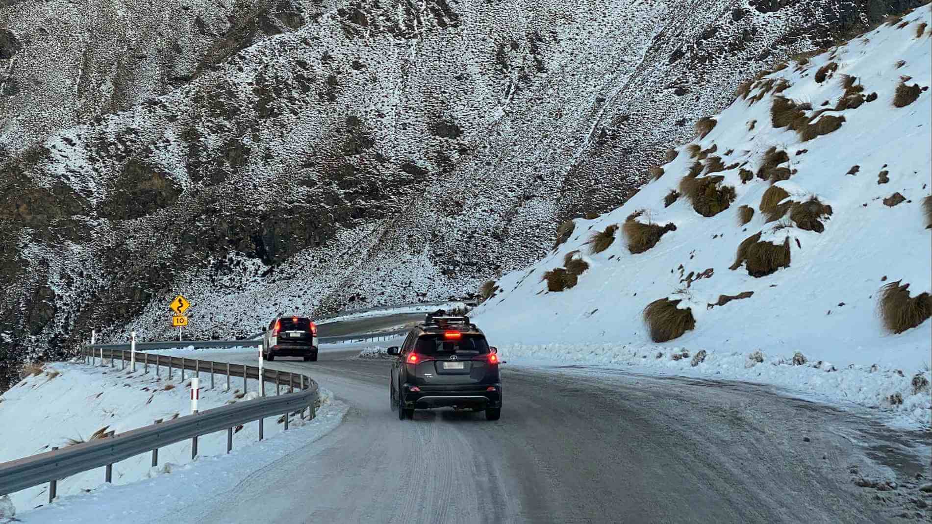 Two SUVs driving on a winding snow-covered mountain road with guardrails and a curve warning sign.