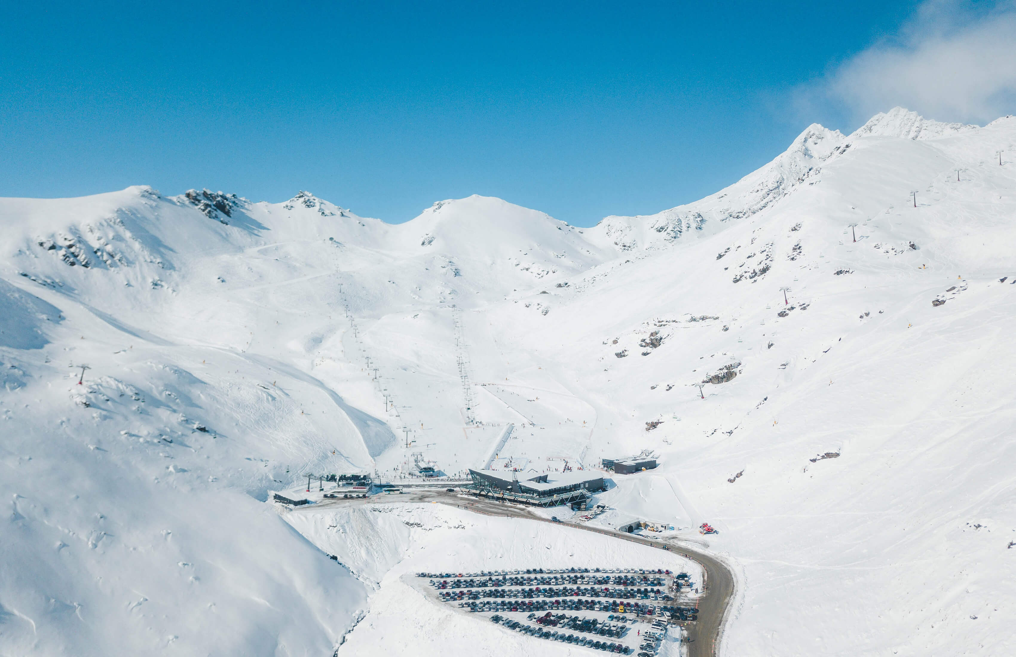 Ski resort with snow-covered mountains, chairlifts, parking lot, and clear blue sky.