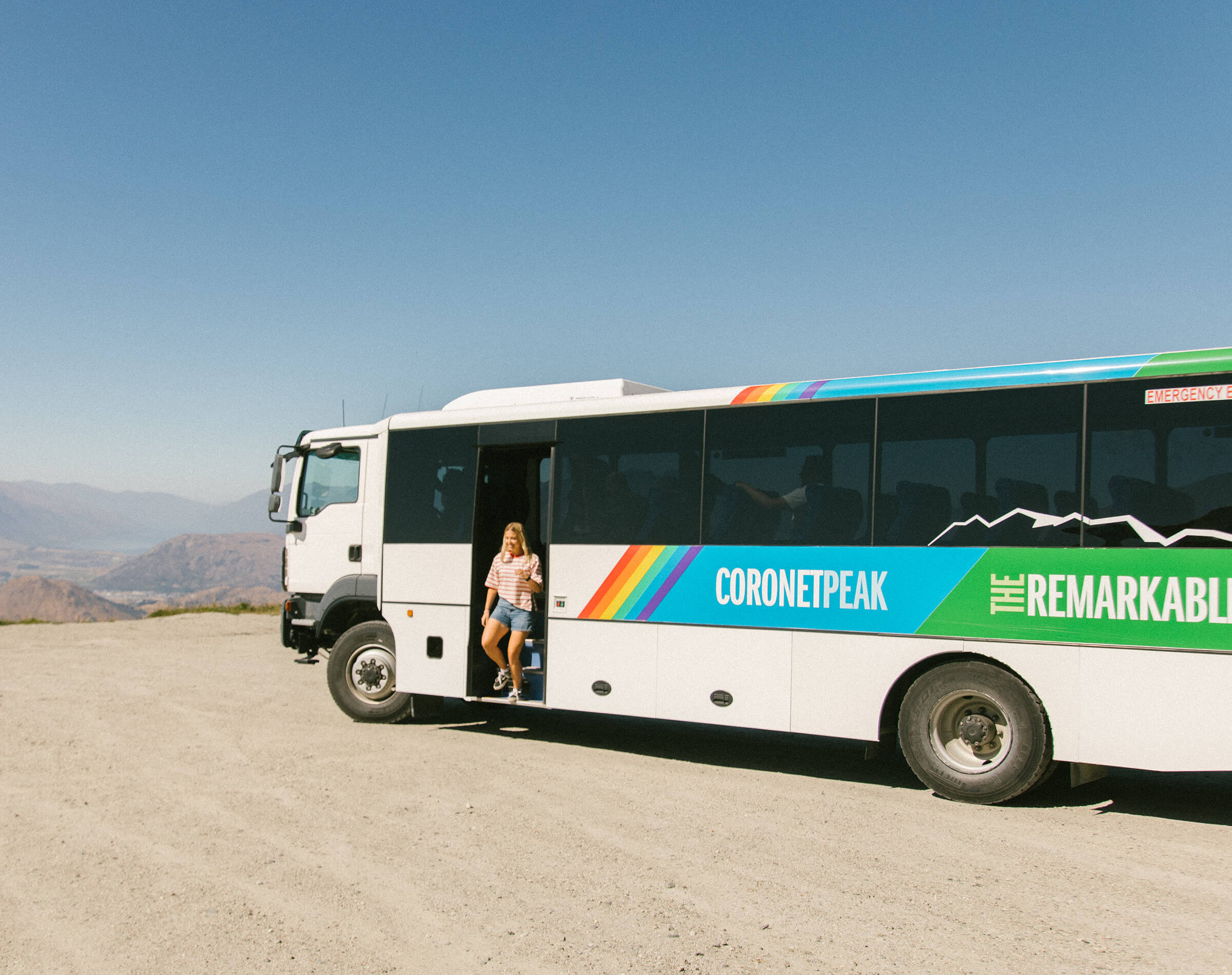 Woman stepping off a white Coronet Peak bus on a sunny day with mountains in the background.