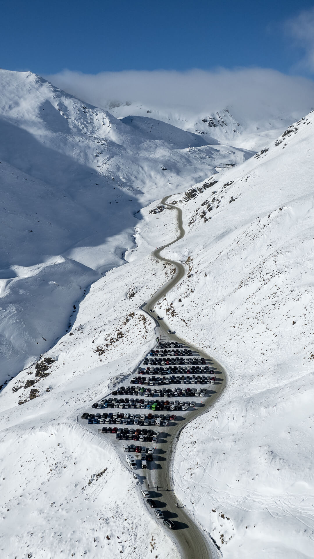 Winding road cutting through snowy mountains with a large parking lot filled with cars.