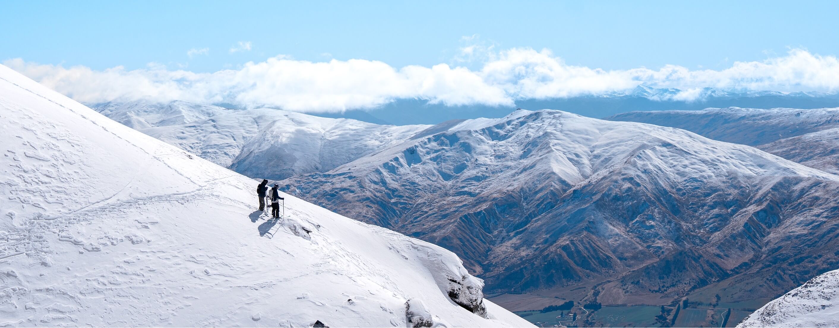 Two skiers pause on a snowy mountain slope with snow-covered peaks and clouds in the distance.