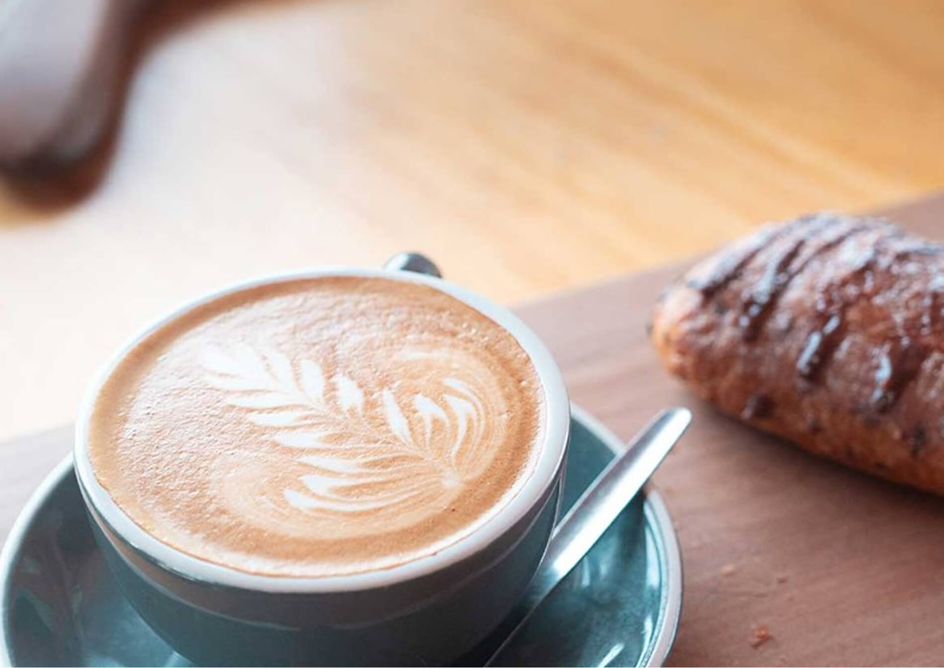 Cup of latte with leaf latte art on a saucer with a spoon beside a chocolate pastry on wooden table.