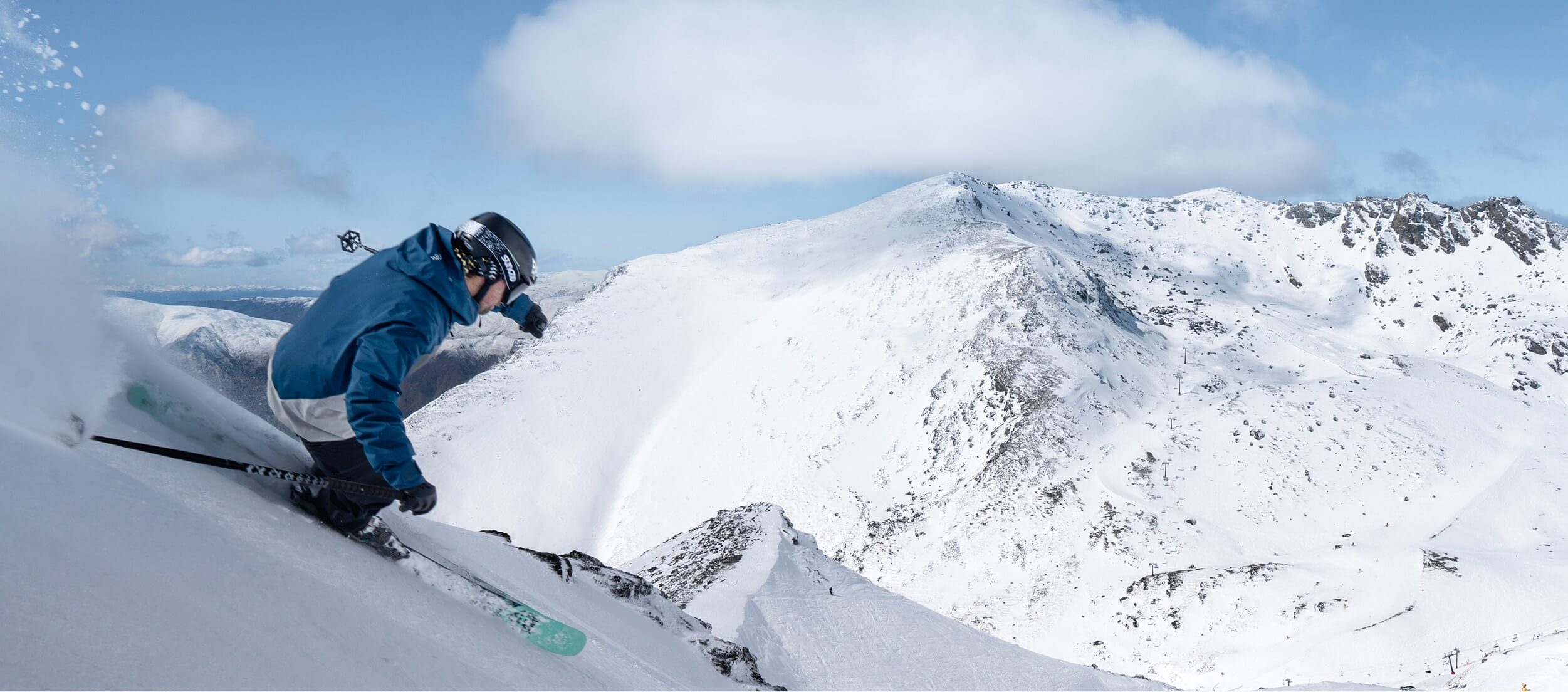 Skier in blue jacket skiing down a snowy mountain slope with snowy peaks and blue sky.