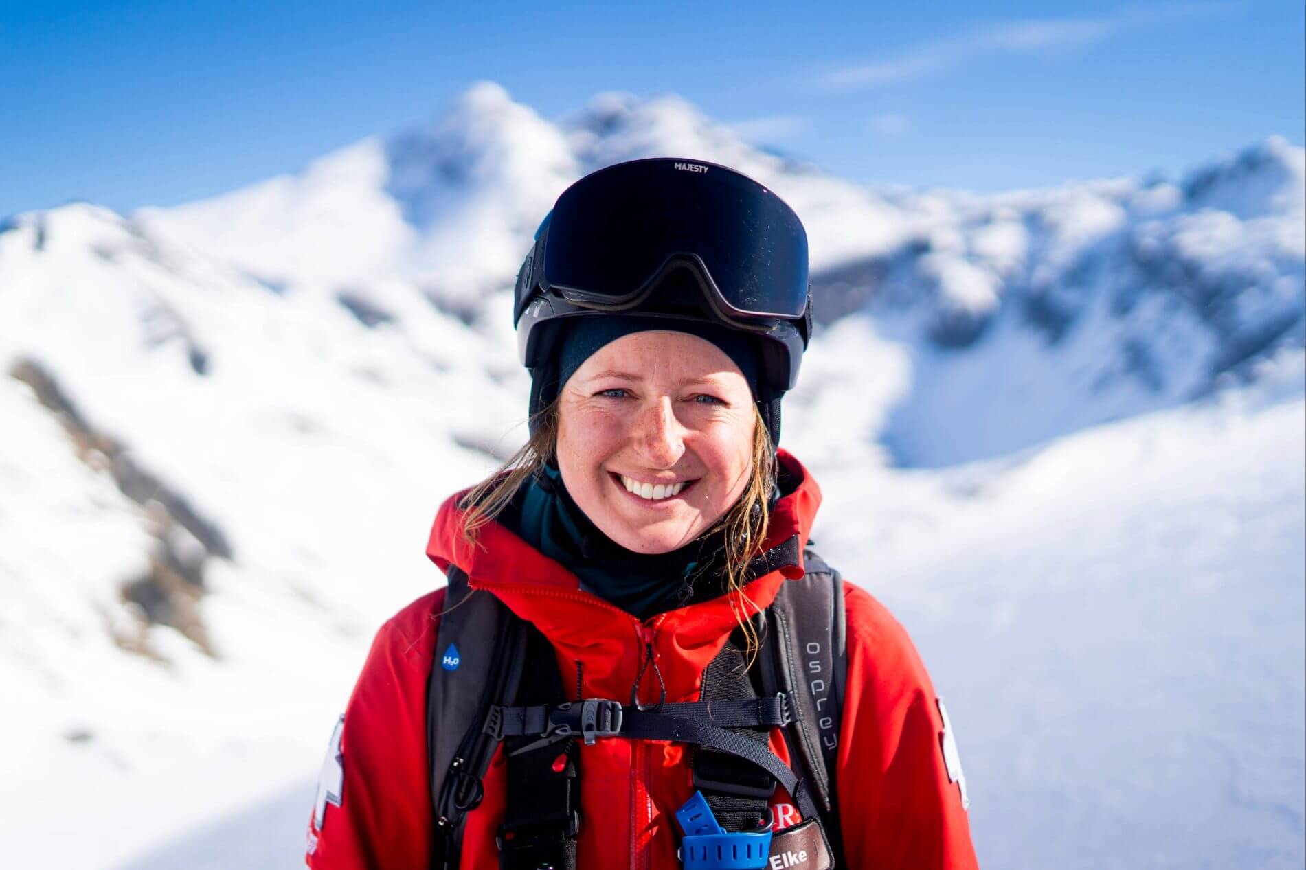 Smiling woman in red ski jacket and helmet with snowy mountain background.
