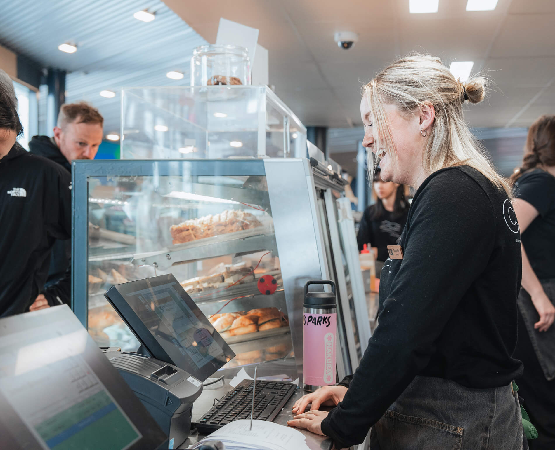 Smiling cafe worker standing behind counter with pastry display and computer screens.