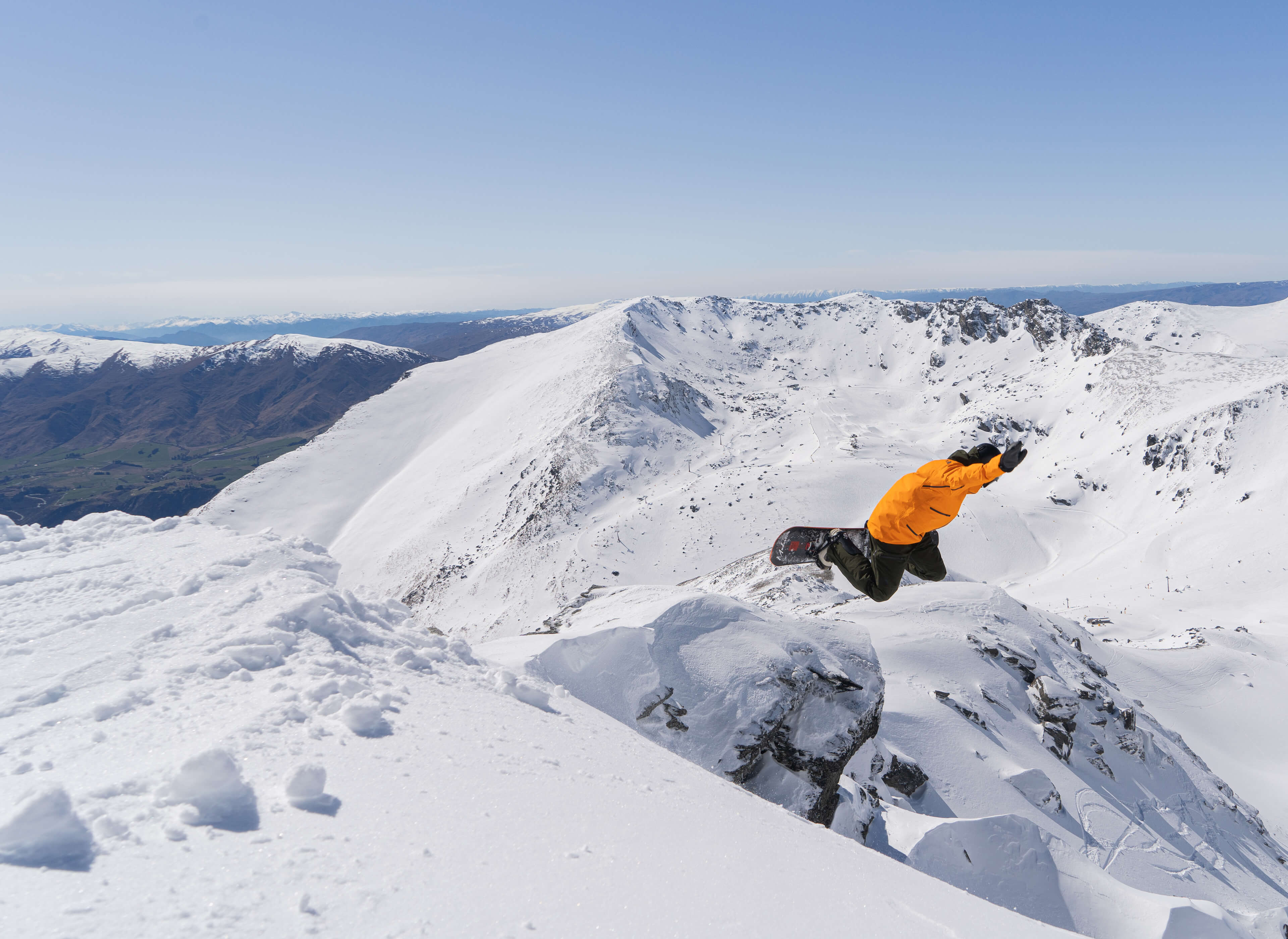 Snowboarder in an orange jacket jumping off a snowy cliff on a bright mountain day.