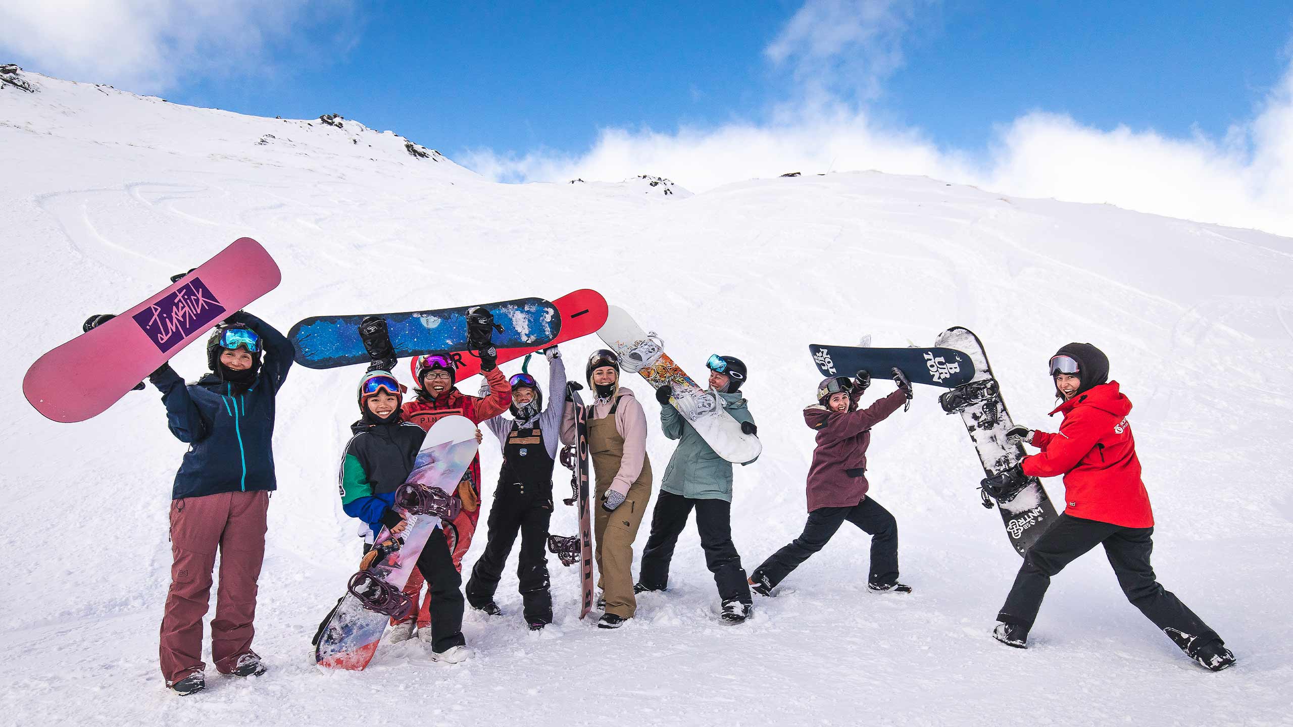 Group of seven snowboarders in colorful winter gear posing with snowboards on snowy mountain slope.