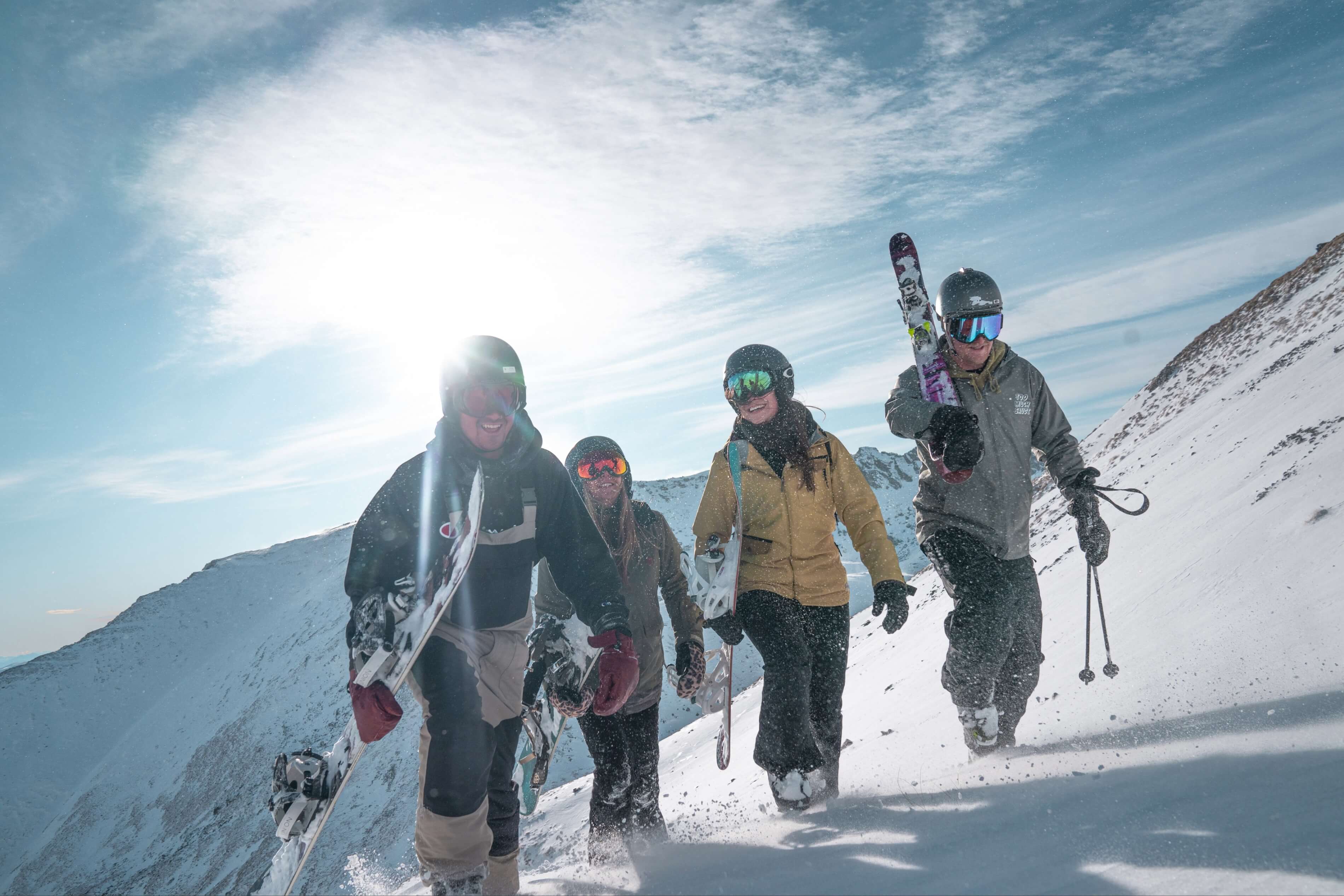 Four skiers and snowboarders walking uphill on snow-covered mountain under blue sky.