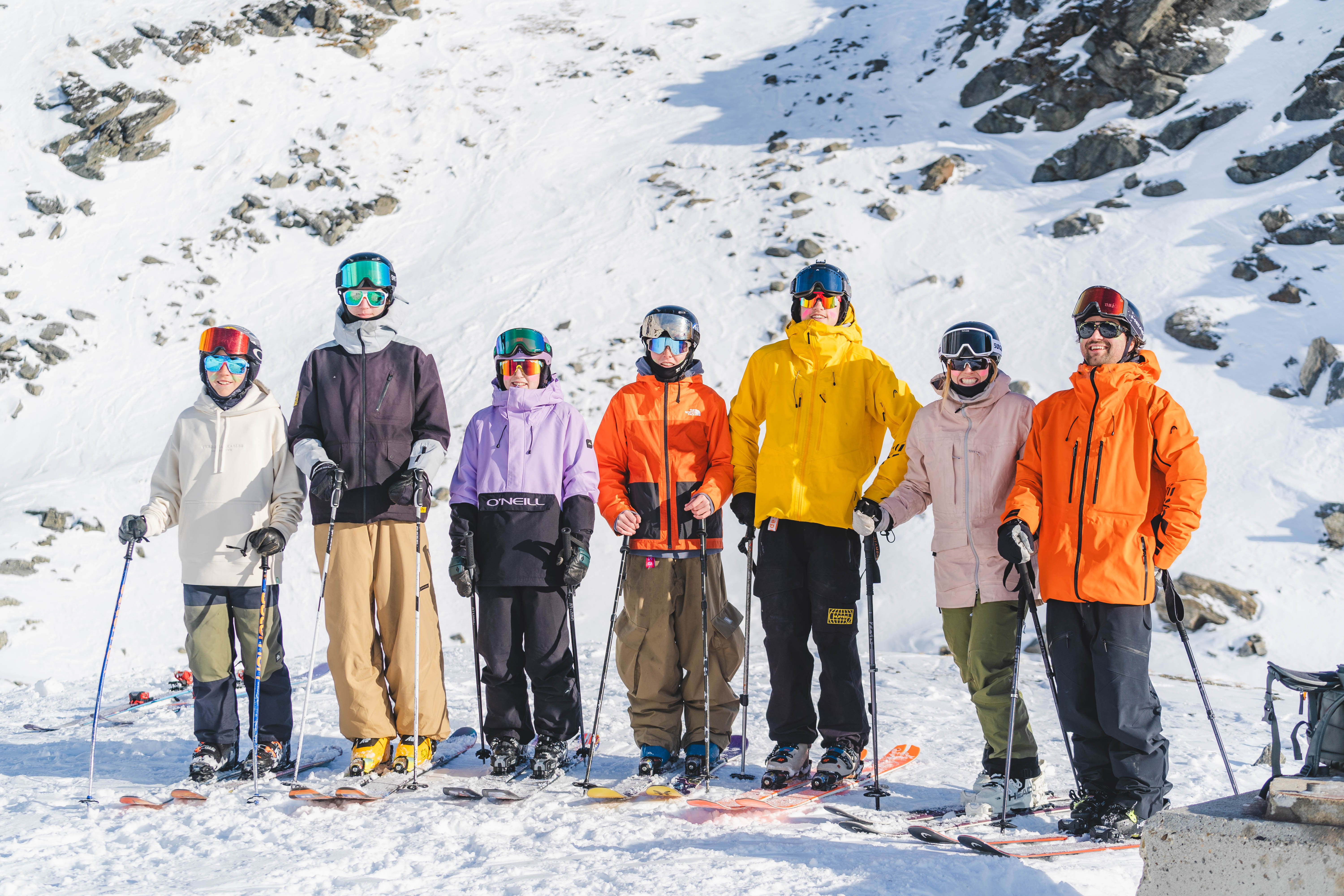 Group of seven skiers in colorful winter gear posing on snow in front of rocky slope on a sunny day.