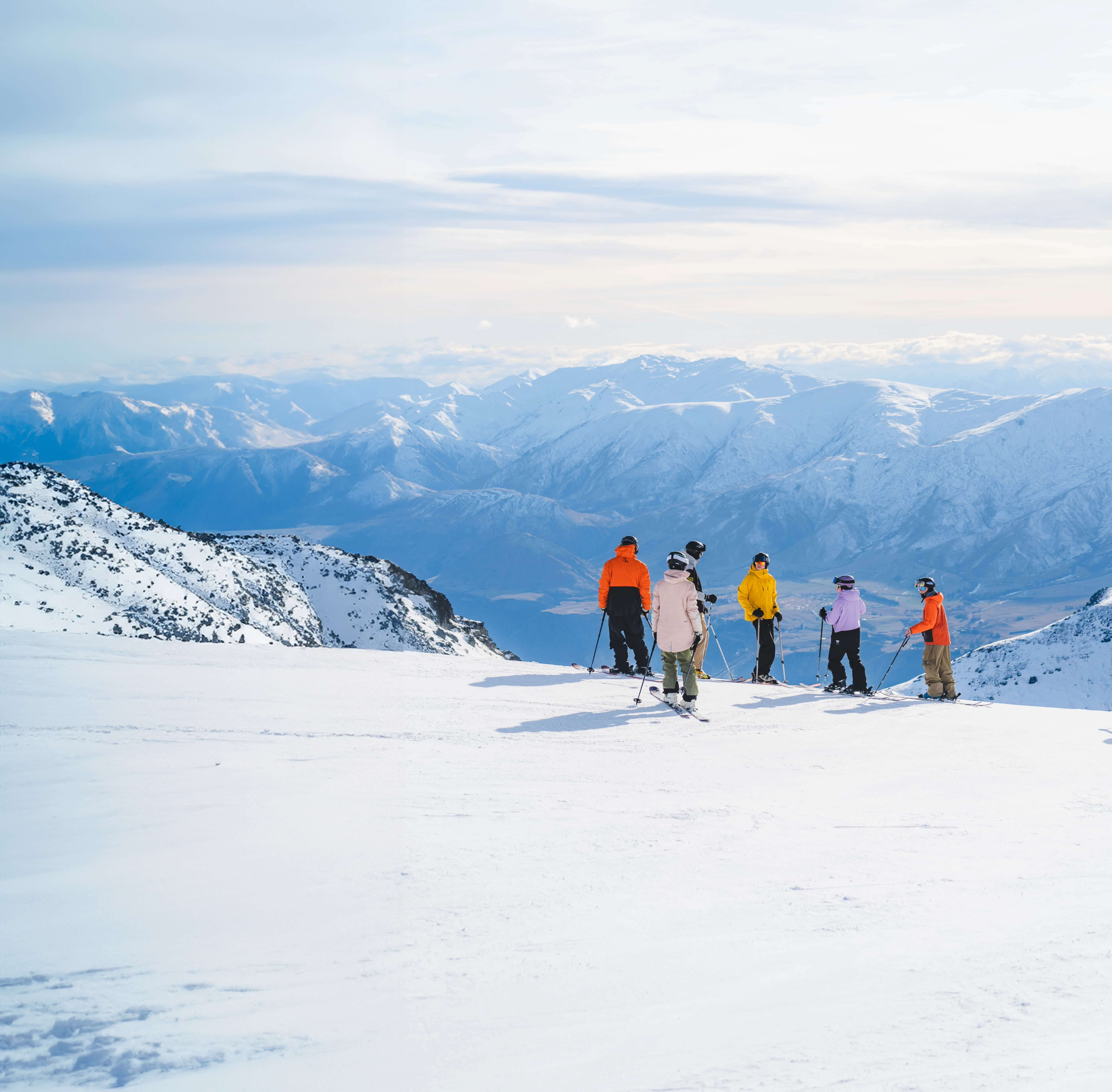 Group of six skiers standing on snow-covered mountain with distant snowy peaks and cloudy sky.