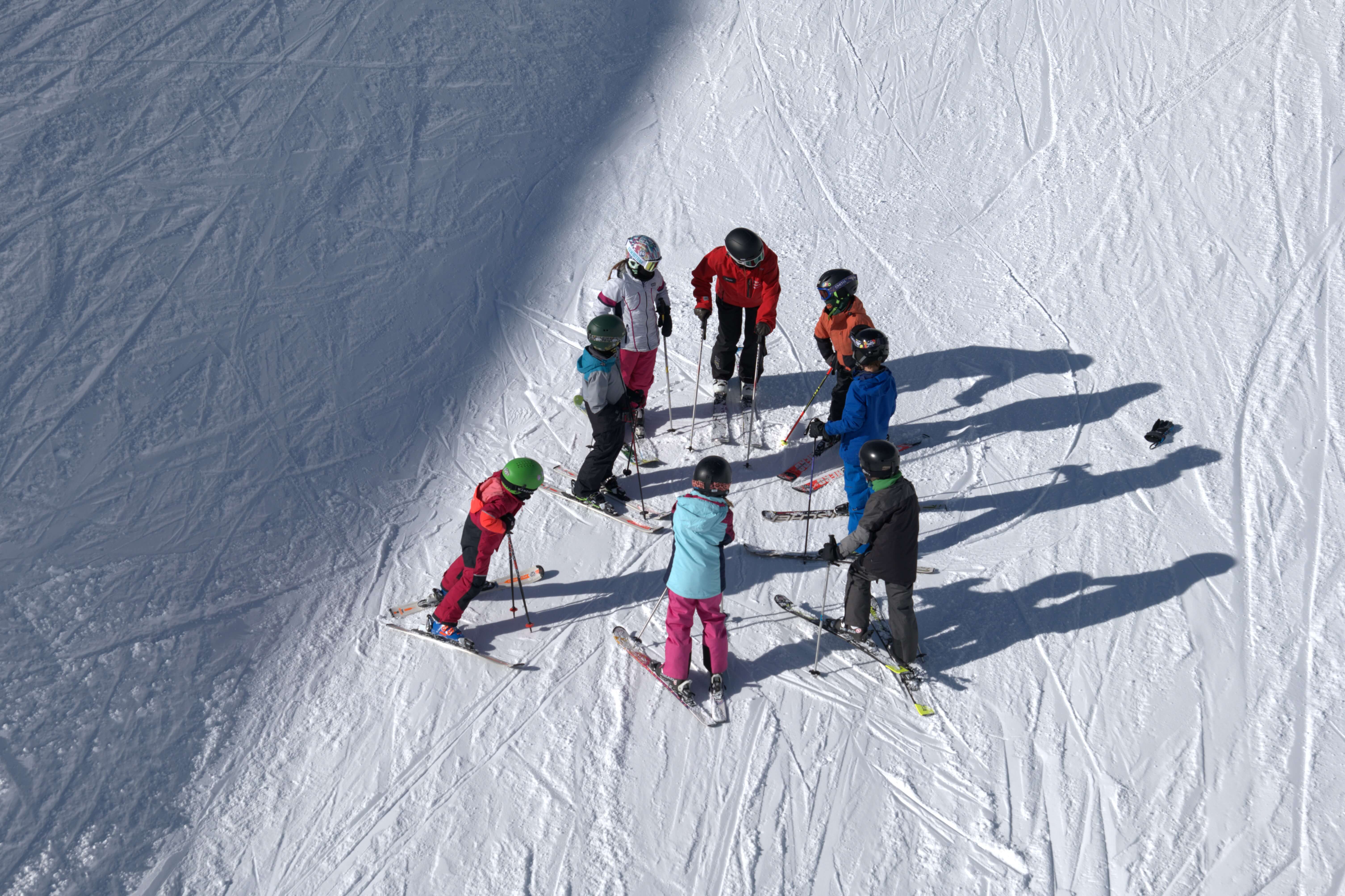 Group of children and instructor in ski gear standing on snowy slope in a circle during lesson.