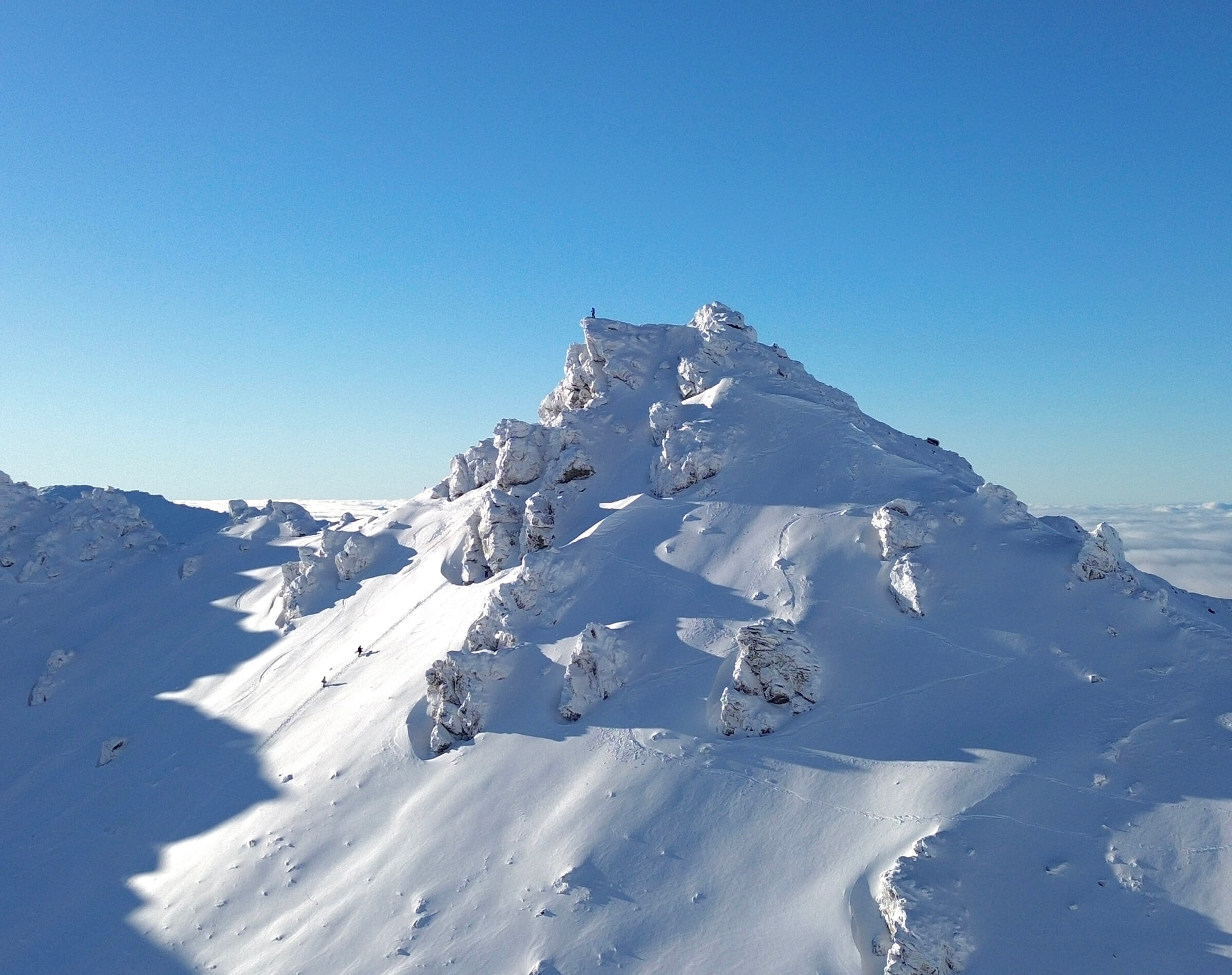 Snow-covered mountain peak under clear blue sky with skiers descending the slope.