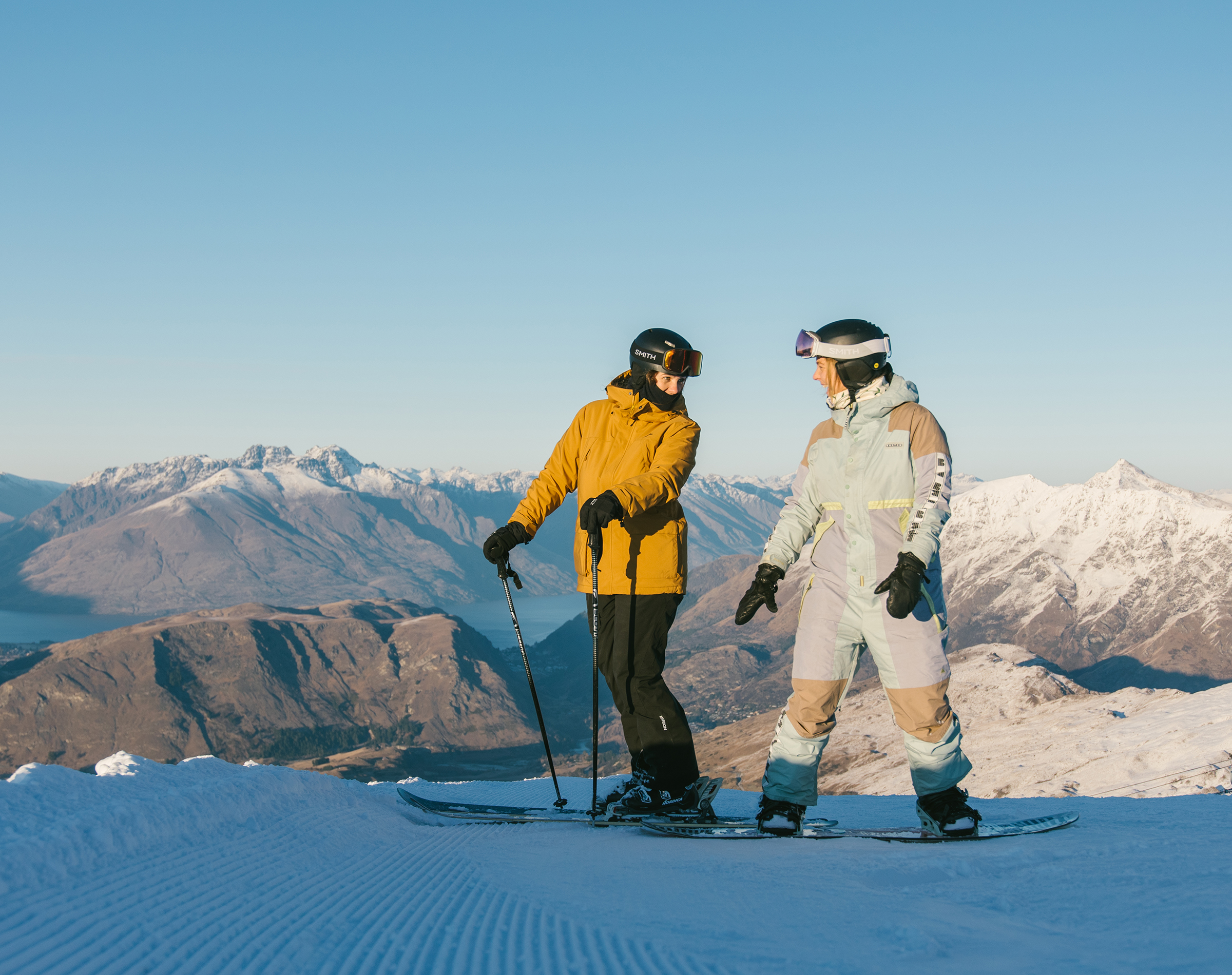 Two people in ski gear standing on a snowy mountain with snowy peaks and clear sky behind.