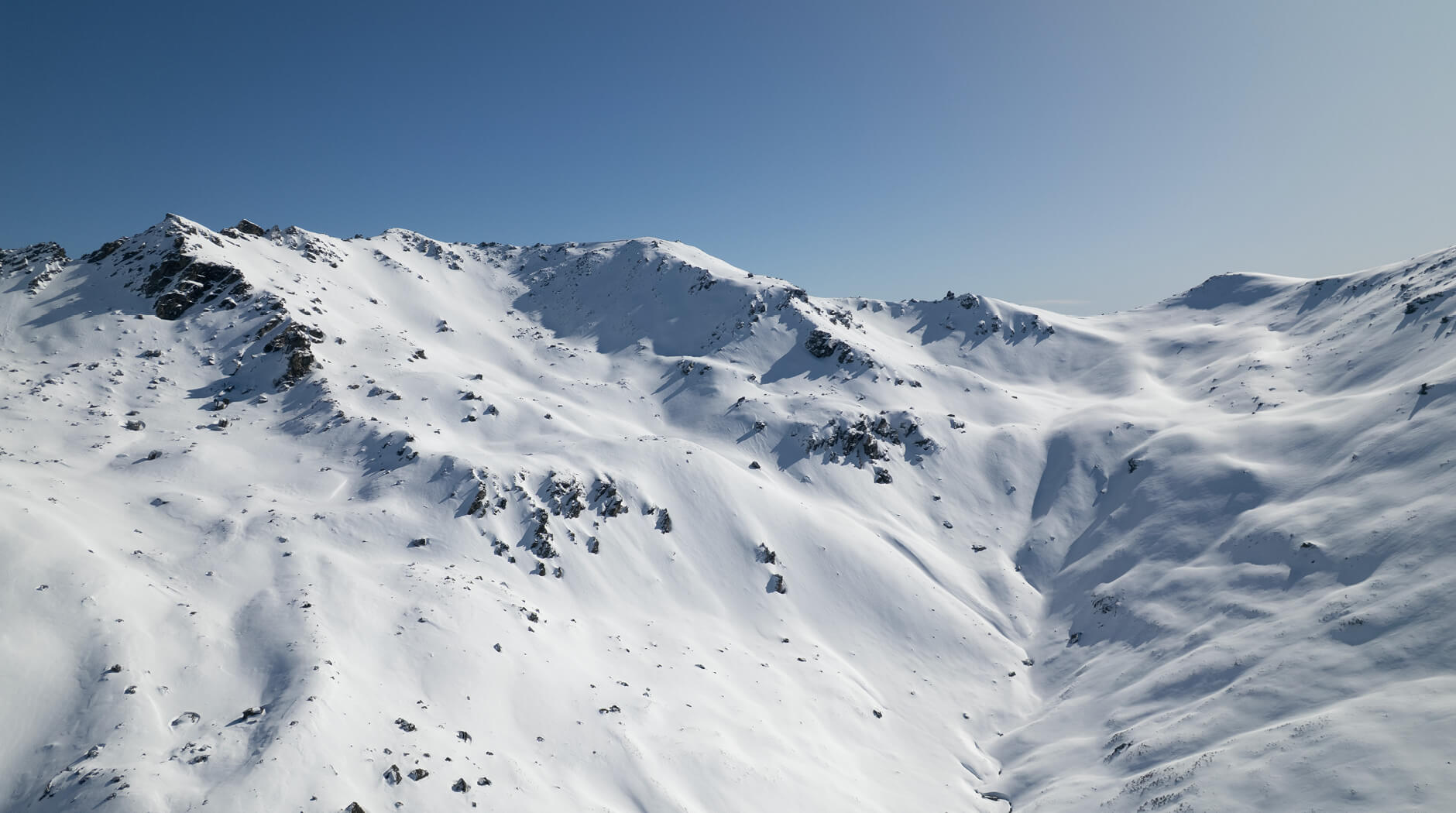 Snow-covered mountain range under clear blue sky with scattered rocks on the slopes.