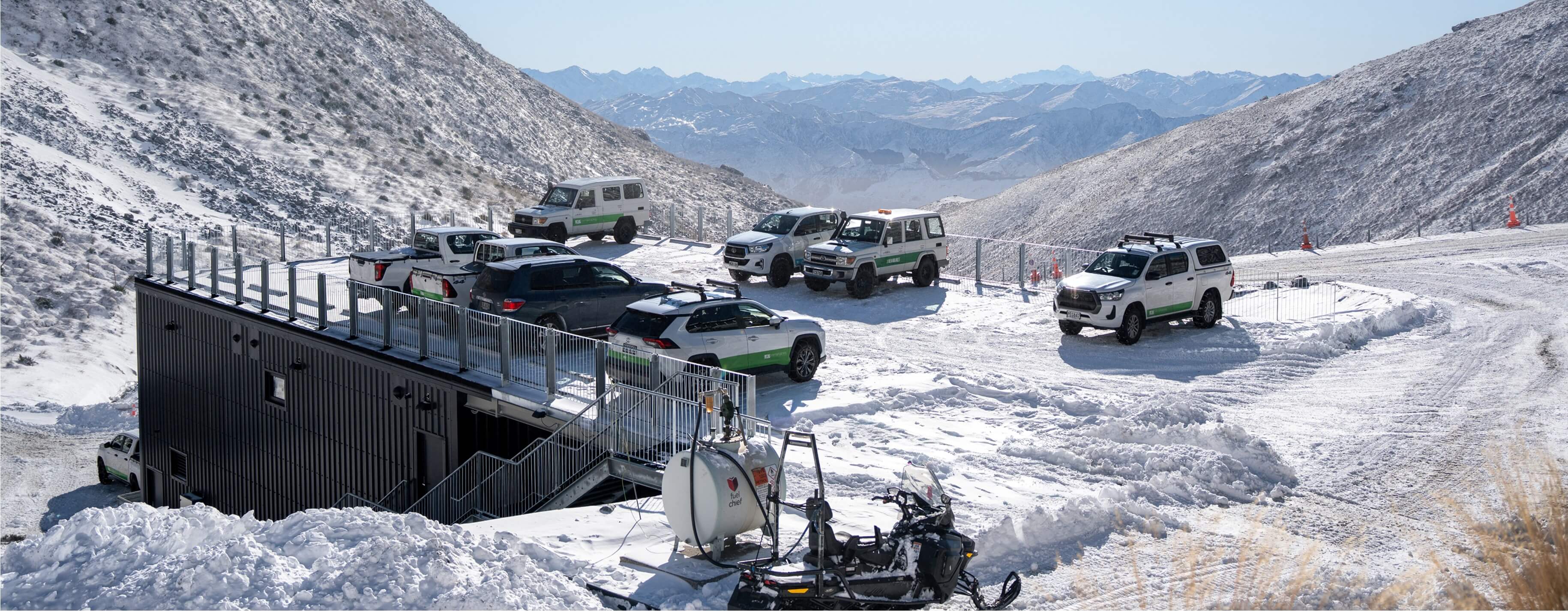 Off-road vehicles parked on a snowy mountain plateau near a black building with metal stairs.