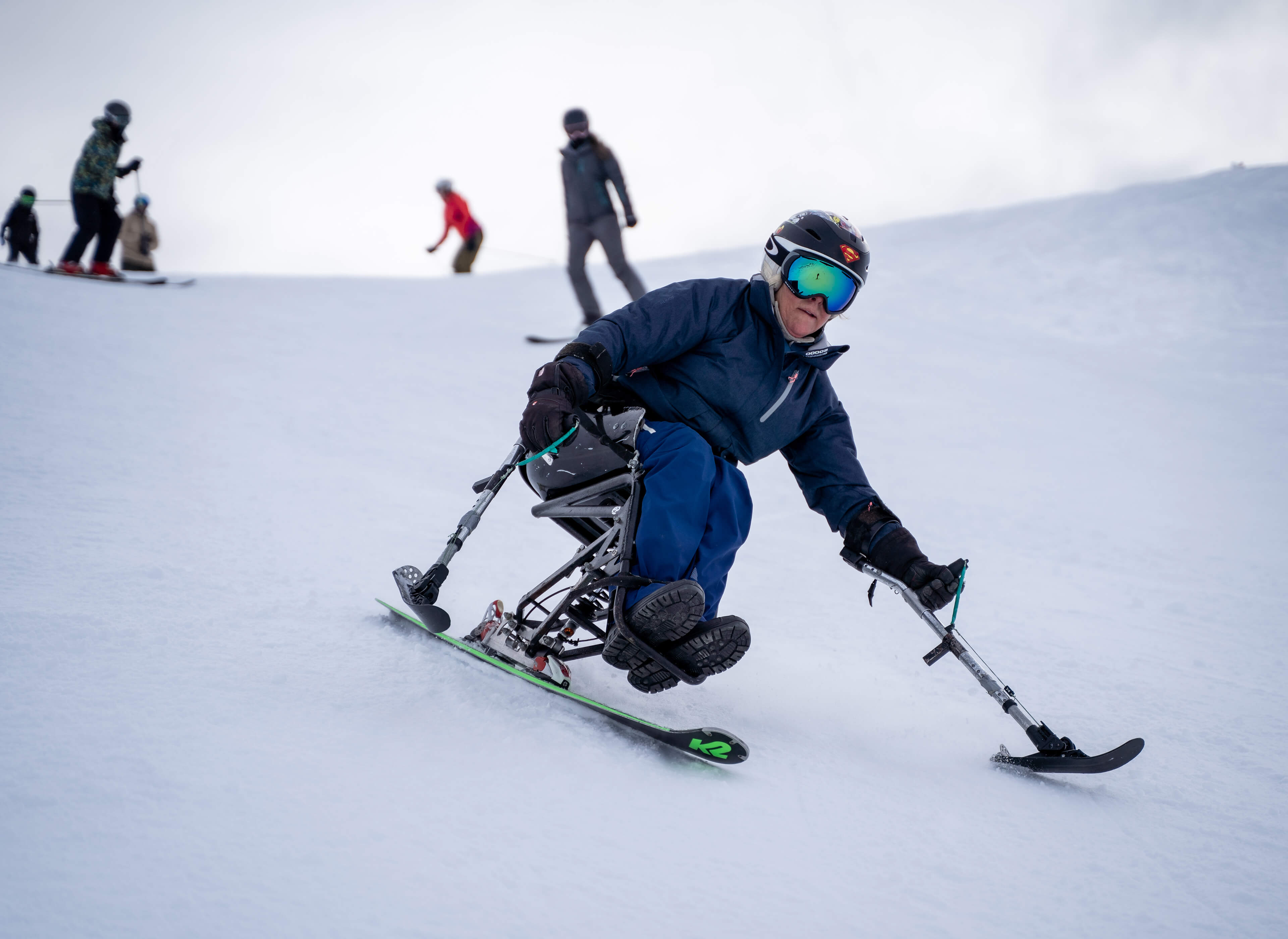 Person in adaptive ski chair skiing down a snowy slope with other skiers in the background.