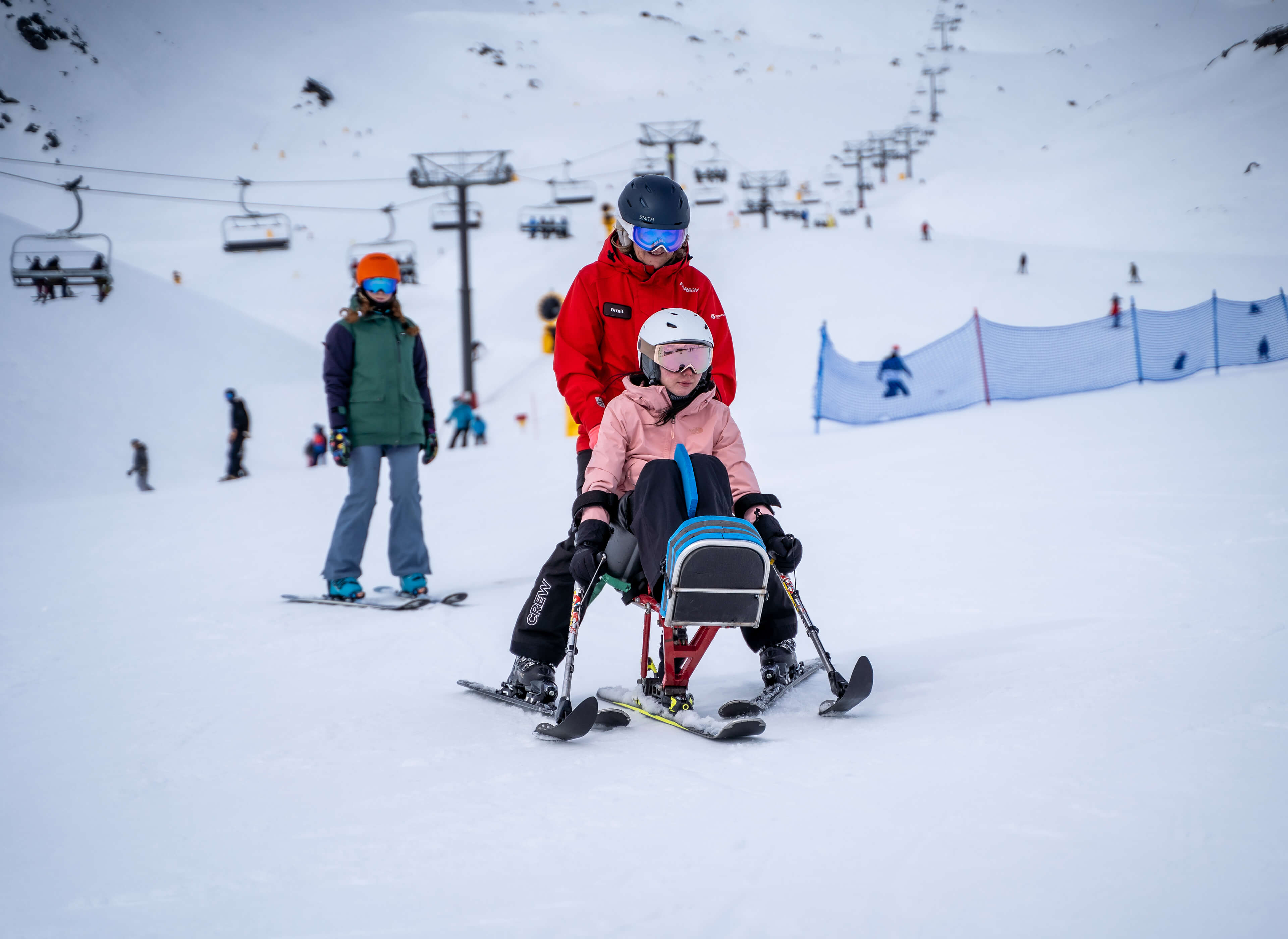 A person in a red jacket assists another in adaptive sit-skiing on a snowy slope with skiers behind.