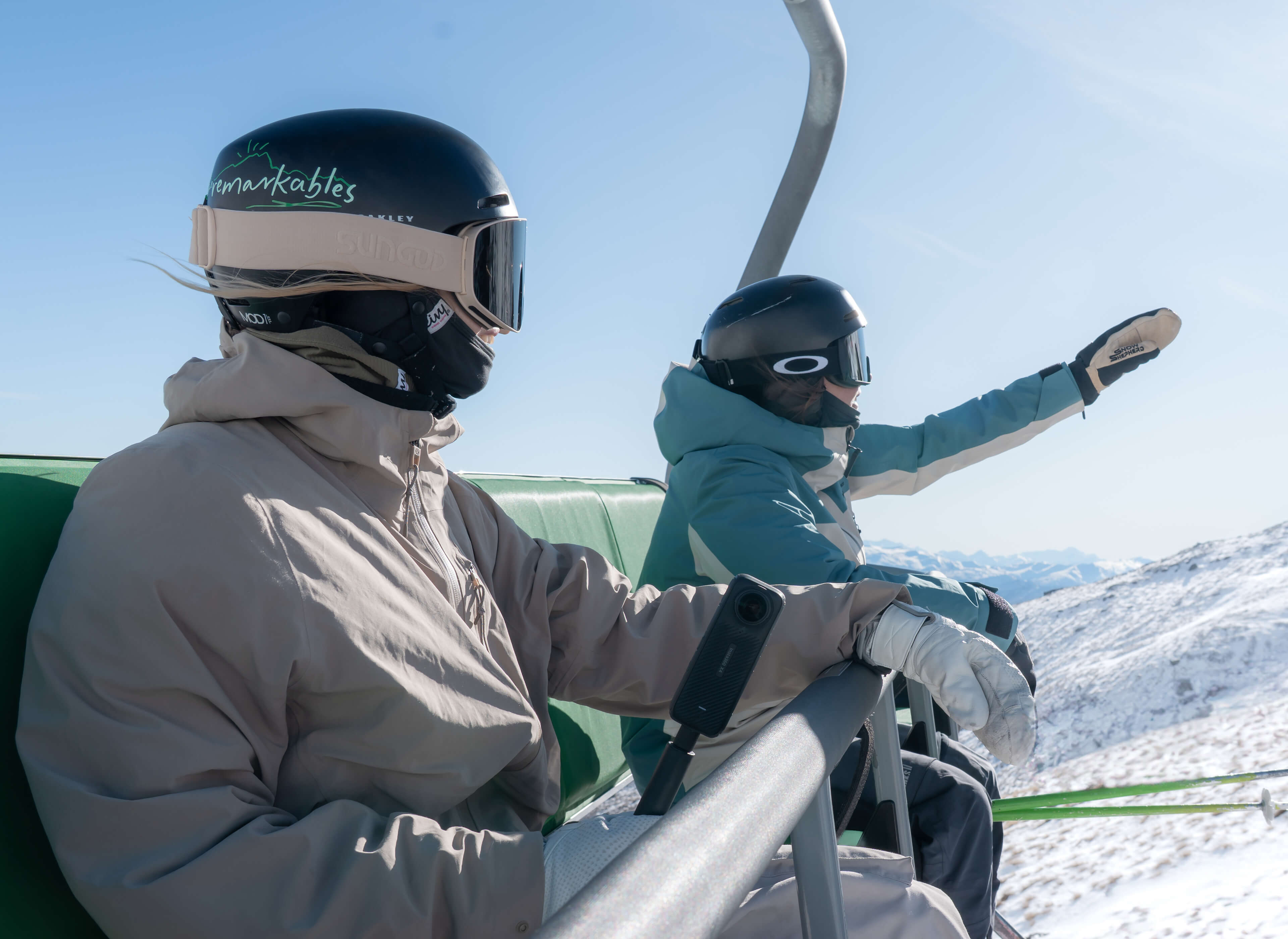Two skiers wearing helmets and goggles riding a ski lift over snowy mountains on a clear day.