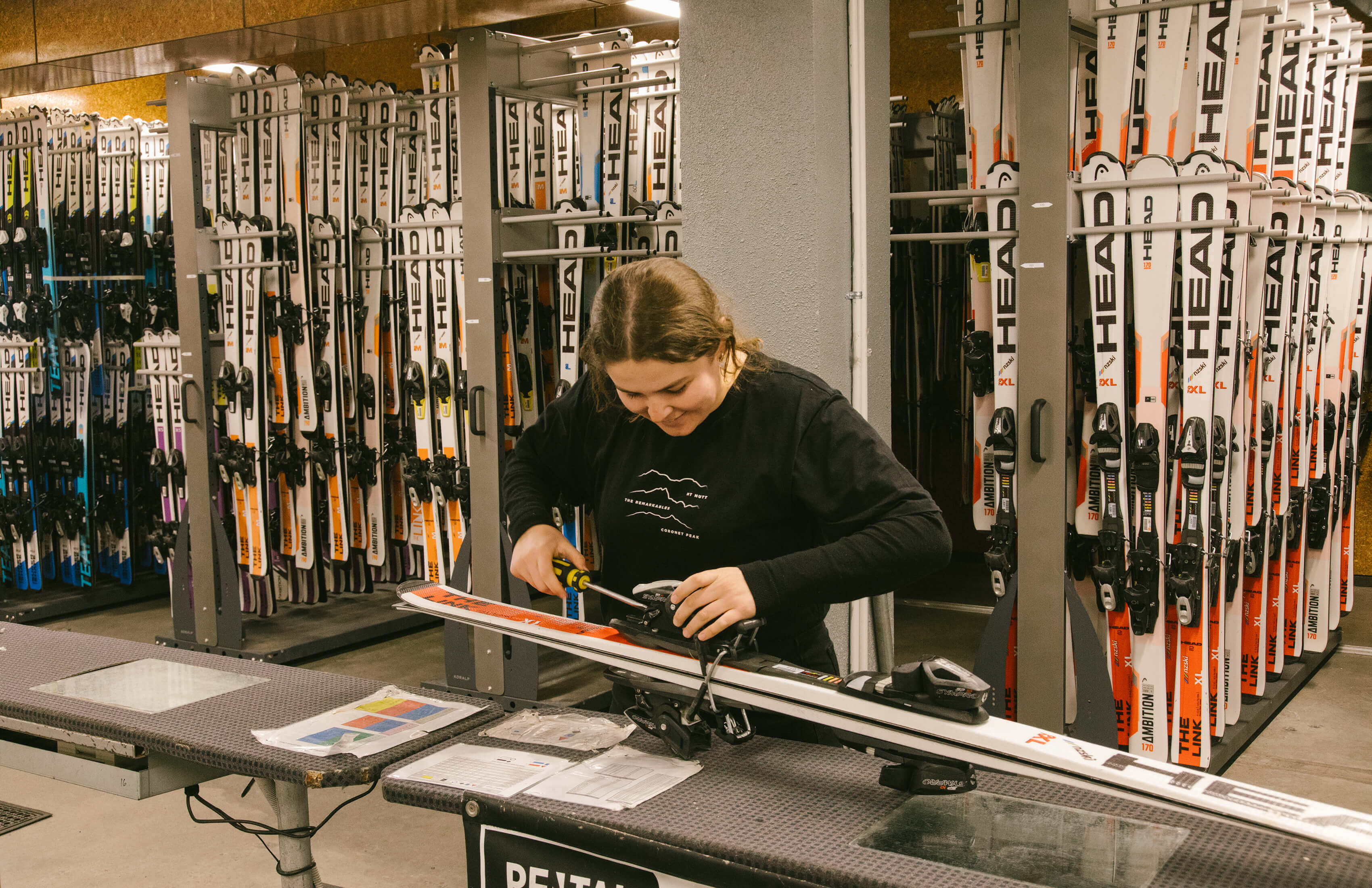 A woman adjusting ski bindings with a screwdriver inside a ski rental shop filled with skis.