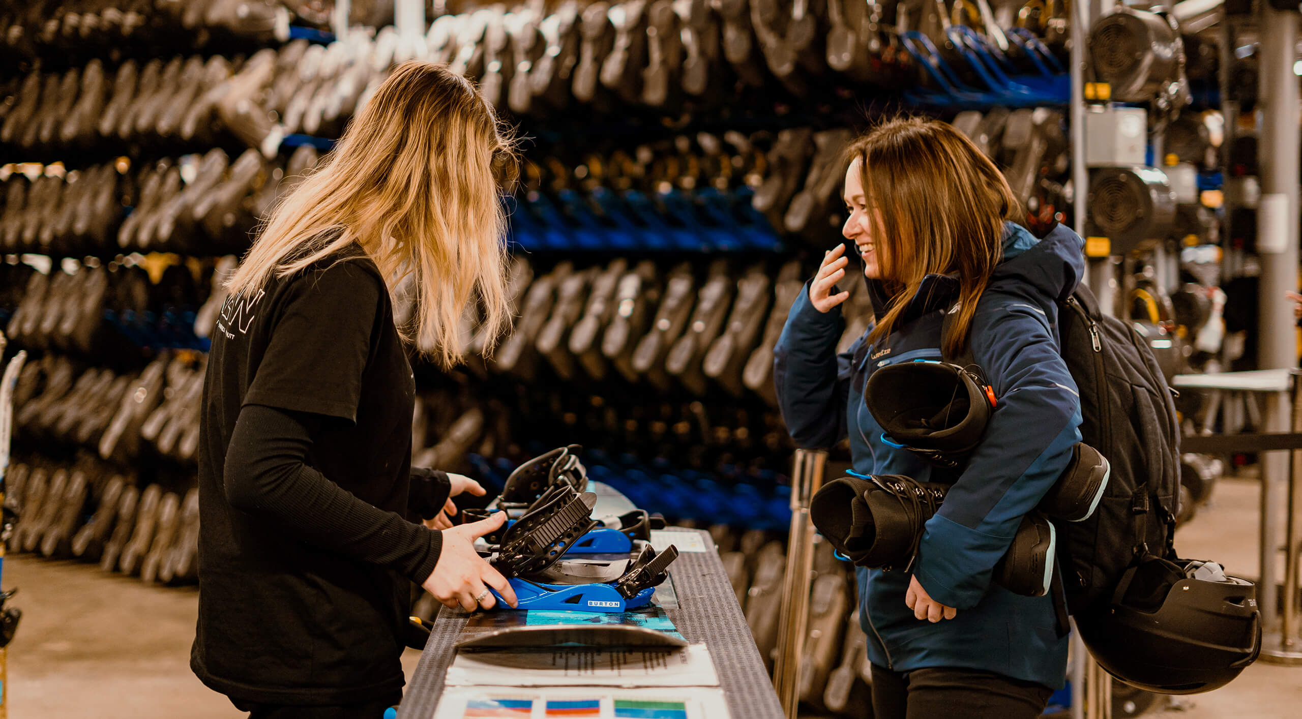 Two women at a rental counter with snowboard gear in a ski rental shop filled with boots.