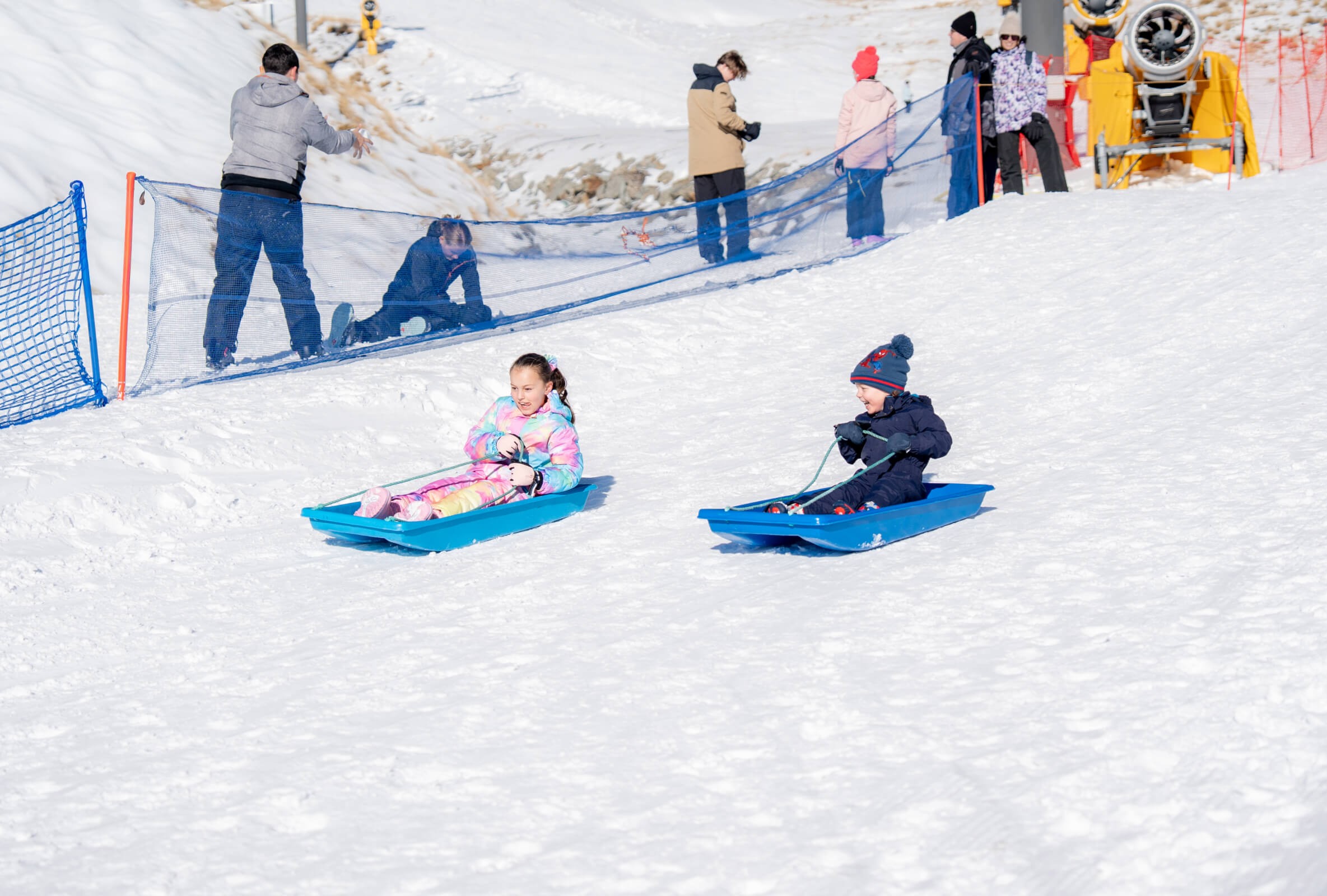 Two children sledding down a snowy hill on blue sleds with people watching behind a safety net.