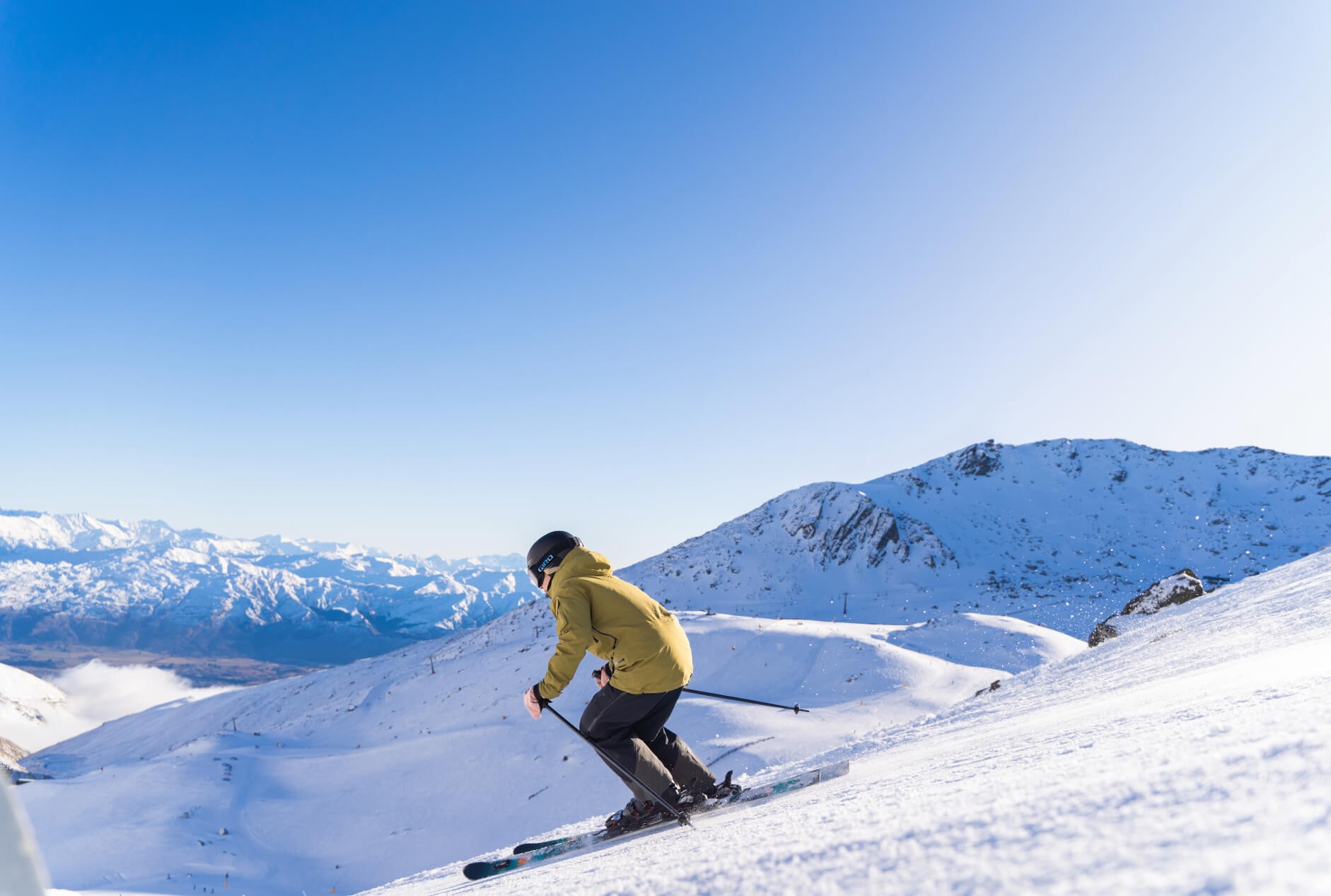 Skier wearing a brown jacket skiing down a ski slope with snow-covered mountains in the background.