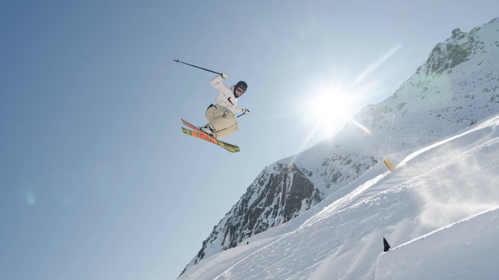 Skier in mid-air performing a jump on a snowy mountain slope with bright sun overhead.