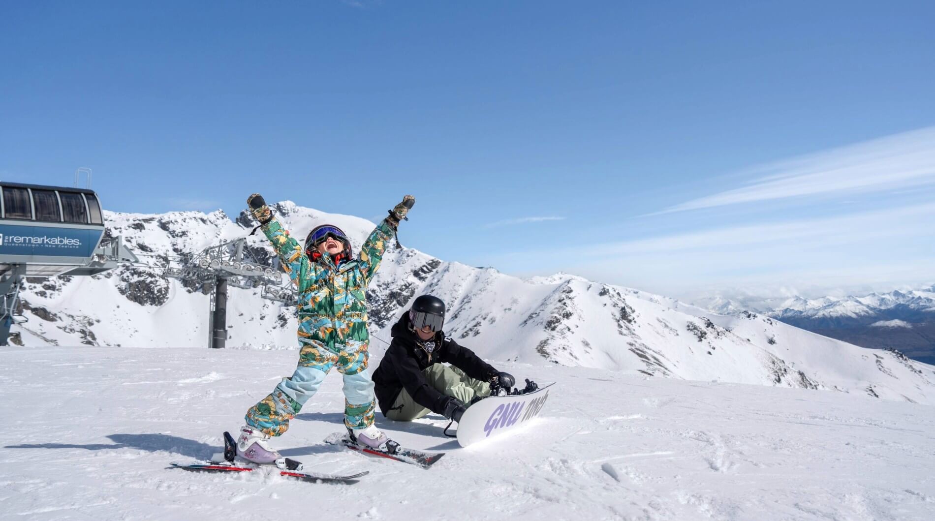Child in colorful ski suit cheering beside seated person with snowboard on snowy mountain under clear sky.