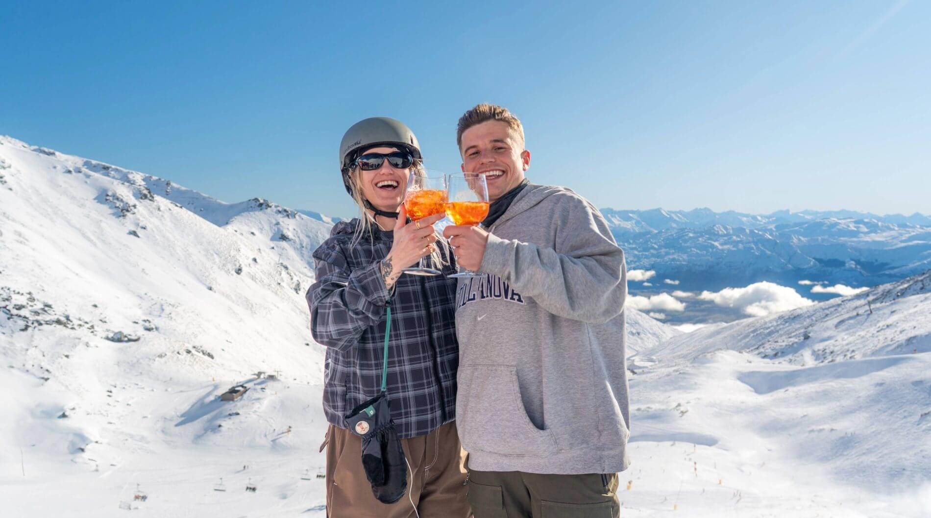 Two smiling people in winter clothes toast with orange drinks on a snowy mountain under blue sky.