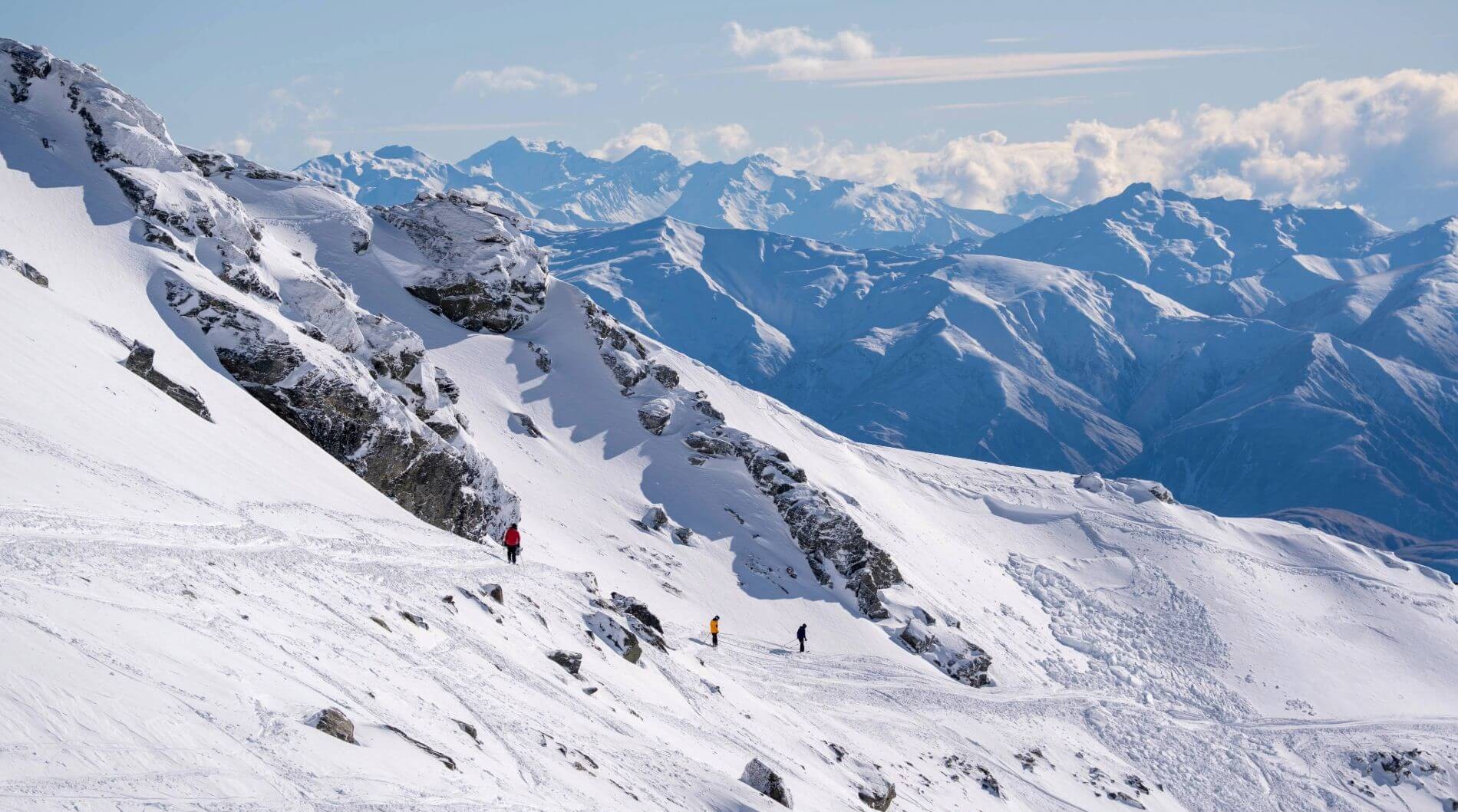 Three skiers in colorful jackets on a snowy mountain slope with rocky outcrops and distant peaks.