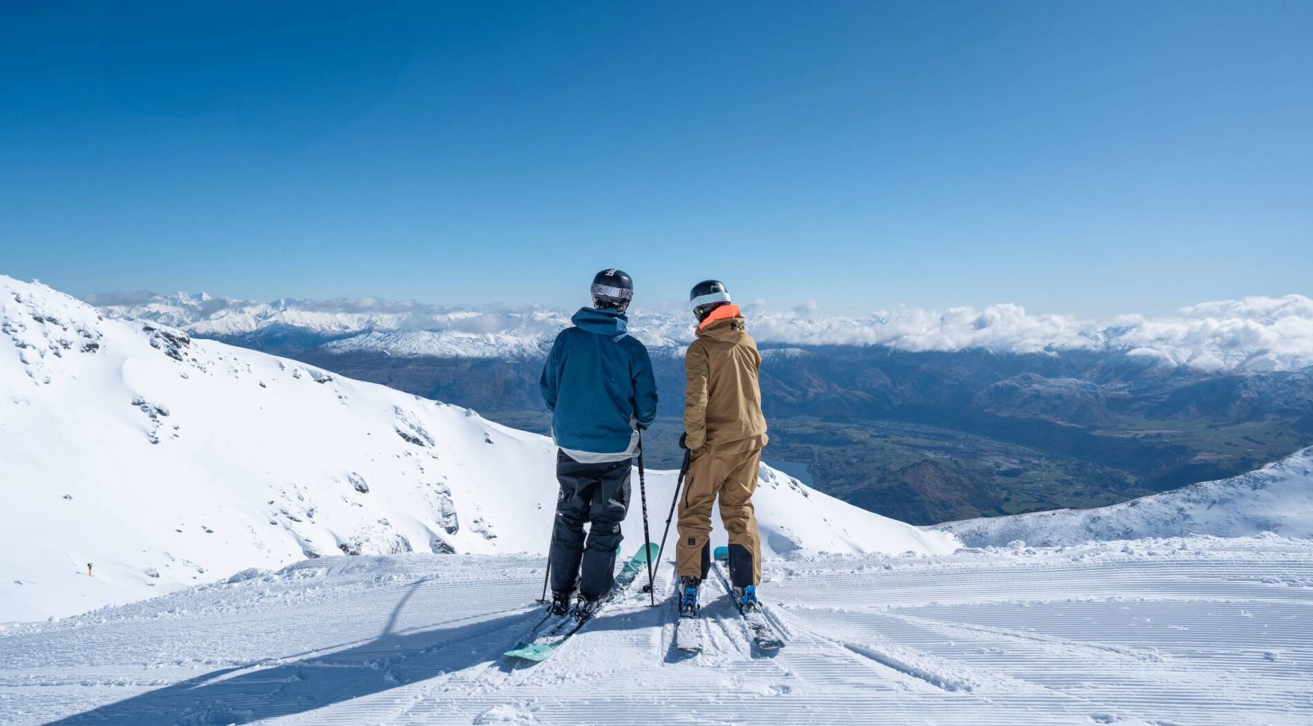 Two skiers standing on snowy slope overlooking distant snow-capped mountains under clear blue sky.