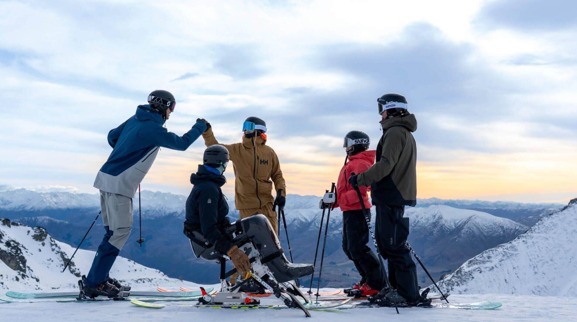 Group of skiers on snowy mountain, one in a sit-ski, celebrating with a fist bump.